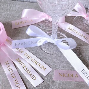 May include: A collection of satin ribbons in light pink and white, featuring gold foil text including names like Rebecca, Francesca, and Nicola, and titles such as Bridesmaid, Maid of Honor, Mother of the Groom, and Bride, tied around glass stems.