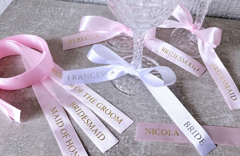 May include: A collection of satin ribbons in light pink and white, featuring gold foil text including names like Rebecca, Francesca, and Nicola, and titles such as Bridesmaid, Maid of Honor, Mother of the Groom, and Bride, tied around glass stems.