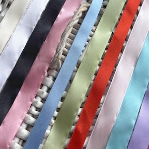 May include: A collection of smooth satin ribbons arranged in a parallel row, featuring a diverse color palette including white, black, pink, light blue, sage green, bright red, lavender, turquoise, and magenta, all with a subtle sheen.