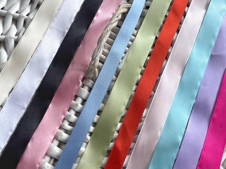 May include: A collection of smooth satin ribbons arranged in a parallel row, featuring a diverse color palette including white, black, pink, light blue, sage green, bright red, lavender, turquoise, and magenta, all with a subtle sheen.