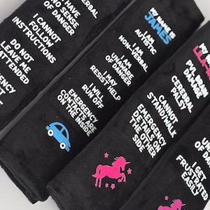 May include: Four black rectangular seat belt covers featuring white text and colorful icons. The text includes personal medical information such as 'I am autistic', 'non-verbal', 'unaware of danger', and 'emergency contact on reverse' with car and unicorn graphics.
