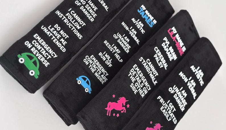 May include: Four black rectangular seat belt covers featuring white text and colorful icons. The text includes personal medical information such as 'I am autistic', 'non-verbal', 'unaware of danger', and 'emergency contact on reverse' with car and unicorn graphics.
