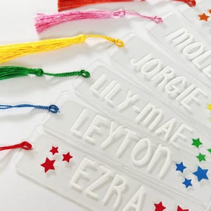 Peut inclure: Une collection de marque-pages rectangulaires en acrylique transparent, chacun portant un nom en relief en lettres blanches. Les noms incluent OLIVIA, MOLLY, JORGIE, LILY-MAE, LEYTON et EZRA. Chaque marque-page est orné de pompons en soie colorés.