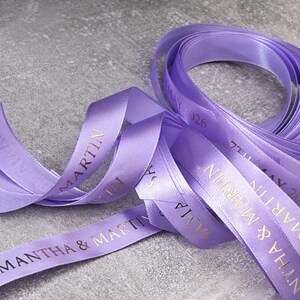 May include: A spool of lavender satin ribbon featuring gold foil text that reads SAMANTHA & MARTIN and 7TH MAY 2026. The smooth, shiny fabric is coiled and draped, showing the repeating personalized names and date printed in a clear, serif font.