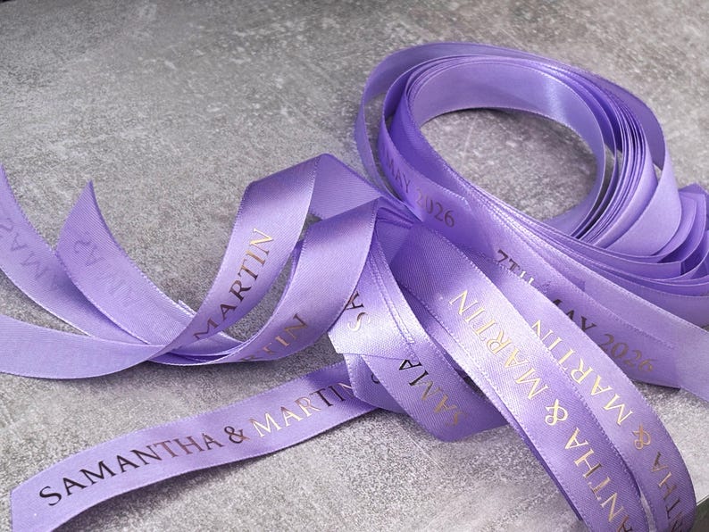 May include: A spool of lavender satin ribbon featuring gold foil text that reads SAMANTHA & MARTIN and 7TH MAY 2026. The smooth, shiny fabric is coiled and draped, showing the repeating personalized names and date printed in a clear, serif font.