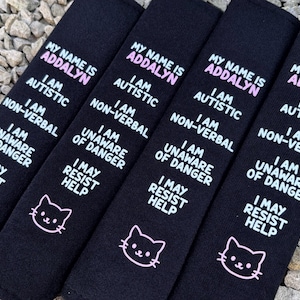 May include: Four black rectangular fabric seatbelt covers featuring white and pink embroidered text that reads: MY NAME IS ADDALYN, I AM AUTISTIC, I AM NON-VERBAL, I AM UNAWARE OF DANGER, I MAY RESIST HELP, and a small white cat face icon.