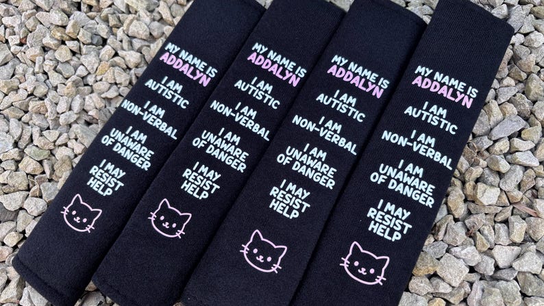 May include: Four black rectangular fabric seatbelt covers featuring white and pink embroidered text that reads: MY NAME IS ADDALYN, I AM AUTISTIC, I AM NON-VERBAL, I AM UNAWARE OF DANGER, I MAY RESIST HELP, and a small white cat face icon.
