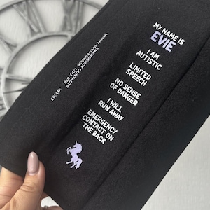 May include: A black rectangular fabric accessory featuring white text that reads: MY NAME IS EVIE, I AM AUTISTIC, LIMITED SPEECH, NO SENSE OF DANGER, I WILL RUN AWAY, EMERGENCY CONTACT ON THE BACK. A small purple unicorn graphic is printed below the text.