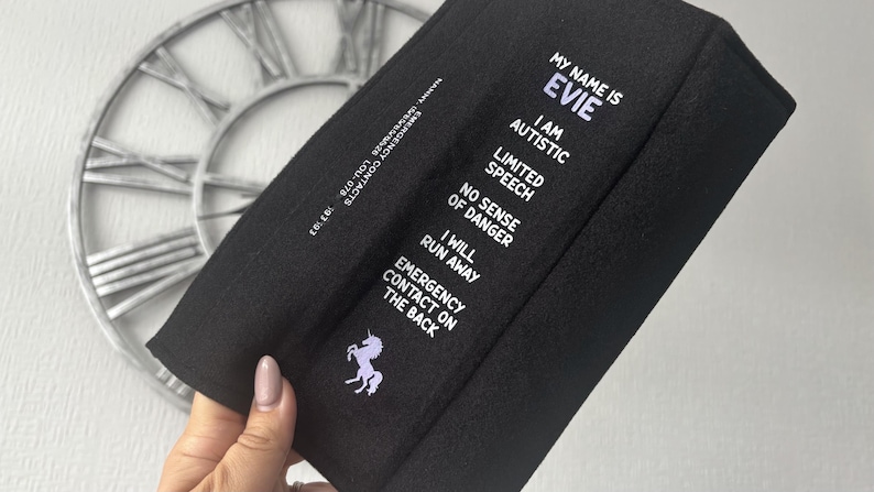 May include: A black rectangular fabric accessory featuring white text that reads: MY NAME IS EVIE, I AM AUTISTIC, LIMITED SPEECH, NO SENSE OF DANGER, I WILL RUN AWAY, EMERGENCY CONTACT ON THE BACK. A small purple unicorn graphic is printed below the text.