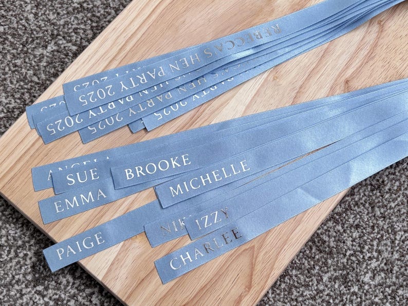 May include: A collection of light blue satin ribbons featuring silver foil text. Some ribbons are printed with the phrase REBECCA'S HEN PARTY 2025, while others are personalized with individual names including SUE, BROOKE, EMMA, MICHELLE, PAIGE, IZZY, and CHARLEE.