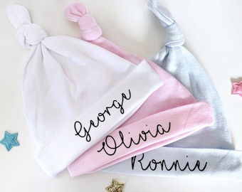 May include: Three knotted baby hats in white, pink, and light blue with the names "George", "Olivia", and "Ronnie" embroidered in black.