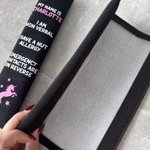 May include: A black fabric seatbelt cover featuring a pink unicorn graphic and white text that reads: MY NAME IS CHARLOTTE, I AM NON VERBAL, HAVE A NUT ALLERGY, EMERGENCY CONTACTS ARE ON REVERSE. The cover has a hook and loop fastener on the interior.