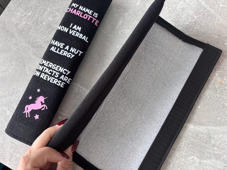 May include: A black fabric seatbelt cover featuring a pink unicorn graphic and white text that reads: MY NAME IS CHARLOTTE, I AM NON VERBAL, HAVE A NUT ALLERGY, EMERGENCY CONTACTS ARE ON REVERSE. The cover has a hook and loop fastener on the interior.