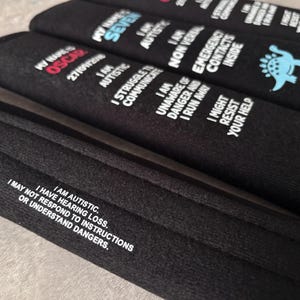 May include: Three black fabric seatbelt covers featuring white printed text. One cover displays 'MY NAME IS OSCAR', 'I AM AUTISTIC', 'I STRUGGLE TO COMMUNICATE', 'I AM UNAWARE OF DANGER AND I RUN AWAY', 'I MIGHT RESIST YOUR HELP'. Another shows a blue dinosaur icon.