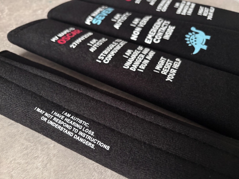 May include: Three black fabric seatbelt covers featuring white printed text. One cover displays 'MY NAME IS OSCAR', 'I AM AUTISTIC', 'I STRUGGLE TO COMMUNICATE', 'I AM UNAWARE OF DANGER AND I RUN AWAY', 'I MIGHT RESIST YOUR HELP'. Another shows a blue dinosaur icon.