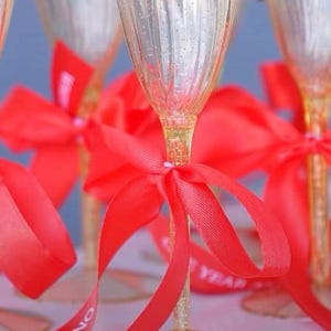 May include: A collection of clear champagne flutes with textured, ribbed bowls and stems filled with gold glitter. Each flute is adorned with a bright red satin ribbon tied in a bow around the stem, with some ribbons featuring white text.