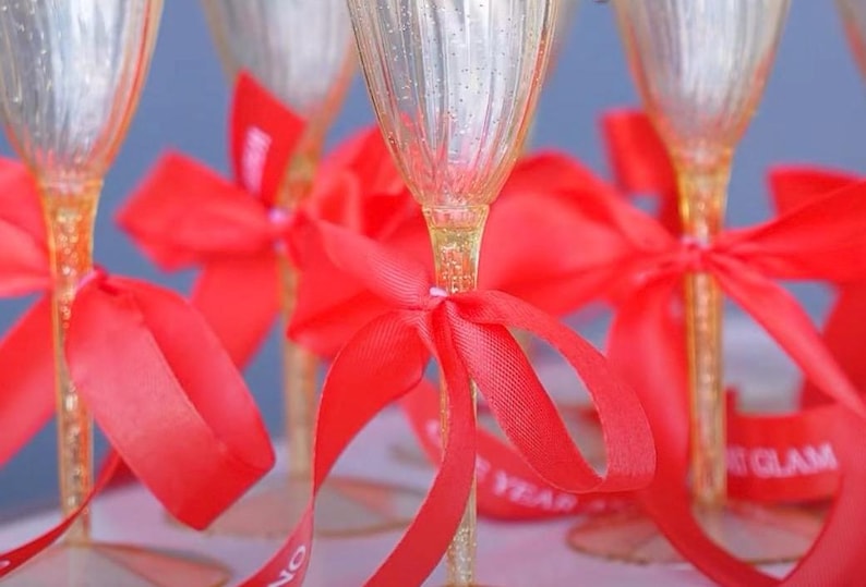 May include: A collection of clear champagne flutes with textured, ribbed bowls and stems filled with gold glitter. Each flute is adorned with a bright red satin ribbon tied in a bow around the stem, with some ribbons featuring white text.