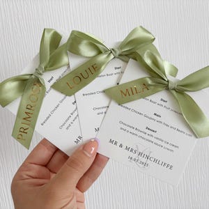 May include: Three white square wedding menu cards with black text, each topped with a sage green satin ribbon bow. The ribbons are printed with gold names: Primrose, Louie, and Mila. The menus list courses and include the text MR & MRS HINCHLIFFE 16.05.2025.
