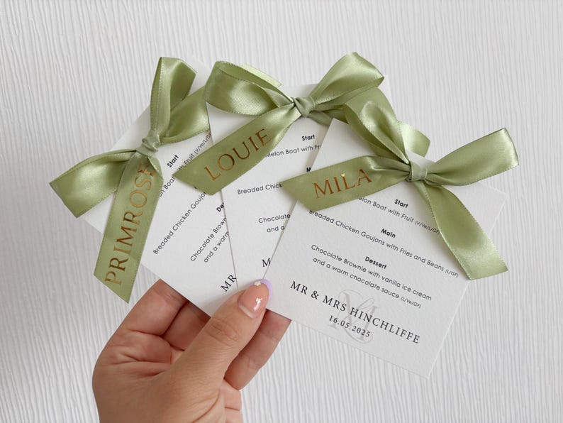 May include: Three white square wedding menu cards with black text, each topped with a sage green satin ribbon bow. The ribbons are printed with gold names: Primrose, Louie, and Mila. The menus list courses and include the text MR & MRS HINCHLIFFE 16.05.2025.