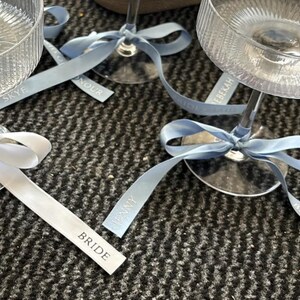 May include: Clear ribbed glass coupes with stems tied with light blue satin ribbons. The ribbons feature printed text including the names Skye, Jenny, and Rebekah, as well as the titles Bride, Maid of Honour, and Bridesmaid in white lettering.