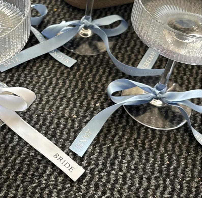 May include: Clear ribbed glass coupes with stems tied with light blue satin ribbons. The ribbons feature printed text including the names Skye, Jenny, and Rebekah, as well as the titles Bride, Maid of Honour, and Bridesmaid in white lettering.