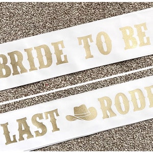 May include: Two white sashes with gold lettering. The top sash says "Bride to Be" with a gold boot icon. The bottom sash says "Last Rodeo" with a gold cowboy hat icon.