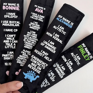 May include: Four black rectangular fabric seatbelt covers featuring white printed text detailing medical conditions and personal information for individuals named Bonnie, Ava, Alexander, and Finley, with small colored icons at the bottom.