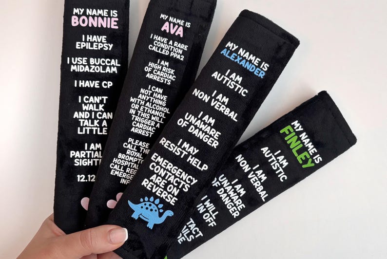 May include: Four black rectangular fabric seatbelt covers featuring white printed text detailing medical conditions and personal information for individuals named Bonnie, Ava, Alexander, and Finley, with small colored icons at the bottom.