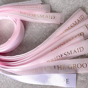 May include: A collection of light pink satin ribbons arranged in a loose coil with several loose ends extending outward. Each ribbon features metallic gold foil text printed on the surface, including the words BRIDESMAID, MOTHER OF THE GROOM, and BRIDE.