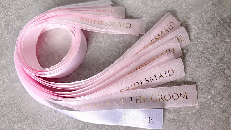 May include: A collection of light pink satin ribbons arranged in a loose coil with several loose ends extending outward. Each ribbon features metallic gold foil text printed on the surface, including the words BRIDESMAID, MOTHER OF THE GROOM, and BRIDE.