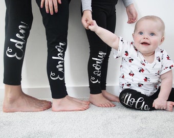 May include: Three children wearing black leggings with silver lettering on the side. The leggings have the names "Eden", "Ember", and "Everly" written on them. A baby is wearing a white t-shirt with a red and white Mickey Mouse print and black leggings with the name "Lola" written on them.