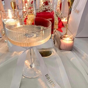 May include: A clear, ribbed glass coupe filled with a sparkling liquid, featuring a white satin ribbon tied around the stem. The ribbon has the word BRIDE printed in gold lettering. The glass is positioned among candles and floral arrangements.