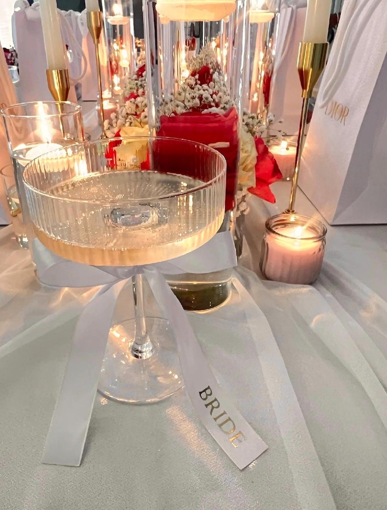 May include: A clear, ribbed glass coupe filled with a sparkling liquid, featuring a white satin ribbon tied around the stem. The ribbon has the word BRIDE printed in gold lettering. The glass is positioned among candles and floral arrangements.