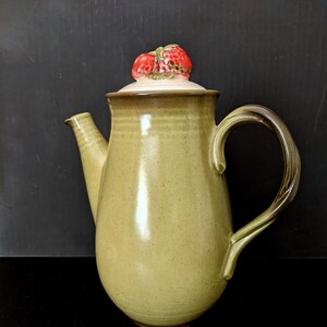 May include: A ceramic teapot with a light green glaze. The lid is topped with a decorative strawberry design. The handle is a twisted design. The teapot has a spout and is likely vintage.