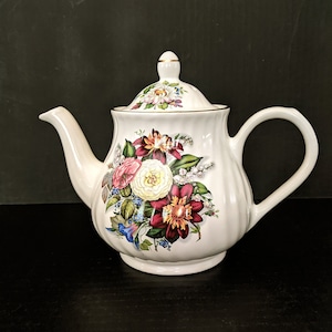 May include: White teapot with a floral design featuring pink, red, yellow, and blue flowers. The teapot has a gold trim around the lid and spout.
