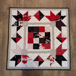 May include: A square quilted wall hanging featuring a central nine-patch block surrounded by star-shaped patterns. The fabric displays a color palette of red, black, white, and cream with floral and geometric prints, finished with a dark patterned border.