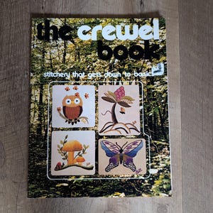 May include: A softcover book titled The Crewel Book: stitchery that gets down to basics. The cover features a forest background with four embroidered squares depicting an owl, a stylized plant, mushrooms with a snail, and a purple butterfly.