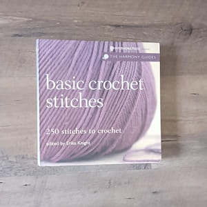 May include: A square book titled Basic Crochet Stitches: 250 stitches to crochet, edited by Erika Knight. The cover features a large, textured ball of light purple yarn. The Interweave Press logo and The Harmony Guides series name are printed at the top.