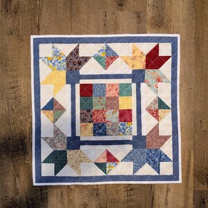 May include: A square patchwork quilt featuring a central grid of sixteen multi-colored floral and patterned fabric squares. The center is framed by blue borders and four corner star motifs made of various printed fabrics on a white background.