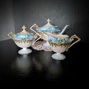 May include: A porcelain tea set consisting of a teapot, a lidded sugar bowl, and a creamer. Each piece features a cream-colored base, a blue floral pattern, and intricate gold filigree detailing along the lower edges and handles.