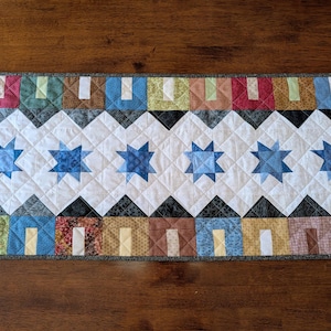 May include: A rectangular quilted table runner featuring a central white band with six blue star patterns. The top and bottom borders consist of a row of small house-shaped fabric blocks in various colors including red, blue, brown, and green.