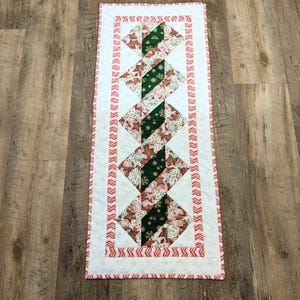 May include: A rectangular Christmas-themed table runner. The runner features a white background with a repeating pattern of red and green diamond shapes. The diamonds are filled with festive patterns, and the edges are trimmed with a red and white candy cane design.