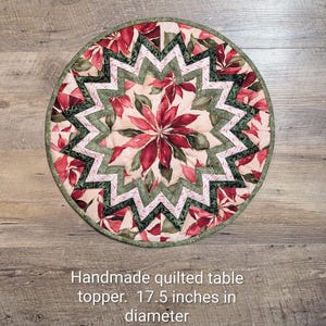 May include: A round quilted table topper measuring 17.5 inches in diameter. The fabric features a central poinsettia floral pattern in shades of red, green, and cream, surrounded by a geometric star design with dark green and white patterned borders.