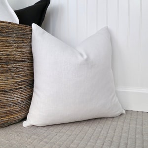 Bright white flax linen pillow cover is stuffed with a feather/down pillow insert and displayed sitting up against a white wall and gray  rug and leaning against a brown woven basket full of similar pillows.