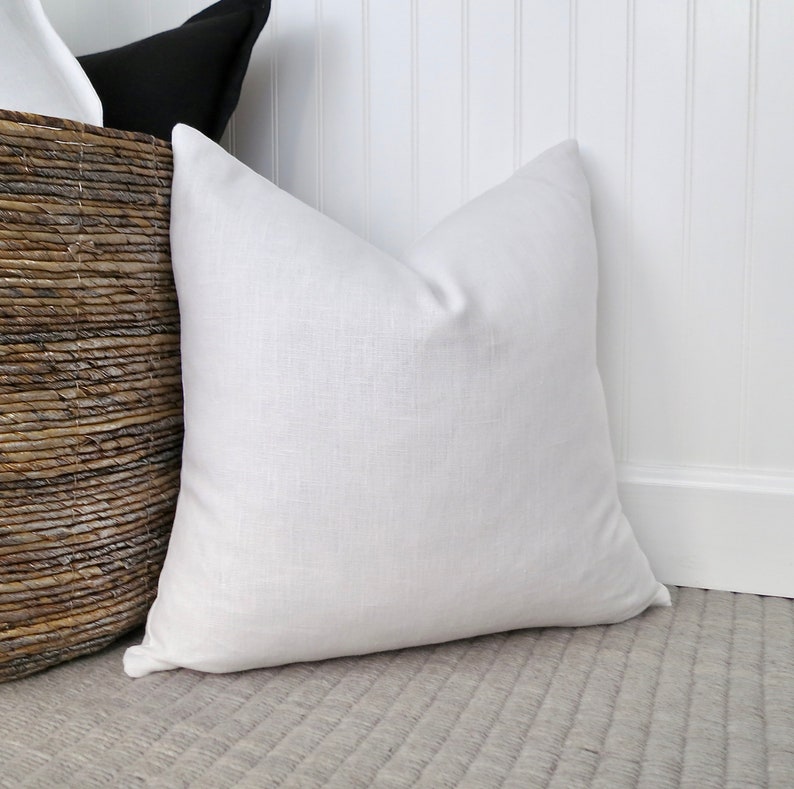 Bright white flax linen pillow cover is stuffed with a feather/down pillow insert and displayed sitting up against a white wall and gray  rug and leaning against a brown woven basket full of similar pillows.