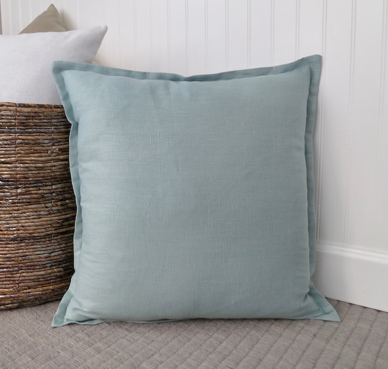 May include: A light blue square throw pillow with a subtle linen texture. The pillow is sitting on a gray carpet.