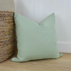 May include: A soft, light green linen pillow with a simple, rectangular shape. The pillow is lying on a light wood floor.