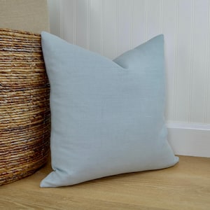 May include: A light blue square pillow made of textured fabric. The pillow is leaning against a white wall and a woven basket. The pillow is a solid color and has a soft, inviting appearance.