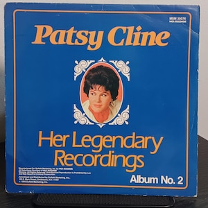 May include: A square vinyl record album cover titled Patsy Cline Her Legendary Recordings Album No. 2. The cover is blue with orange text and features a central oval portrait of Patsy Cline framed by white decorative scrollwork and small text.