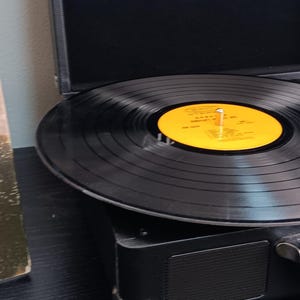 Pode incluir: Um disco de vinil preto tocando em um toca-discos preto vintage. O toca-discos est&aacute; aberto, com a agulha repousando sobre o vinil. Uma capa de &aacute;lbum &eacute; vis&iacute;vel ao fundo. O r&oacute;tulo do disco &eacute; amarelo.